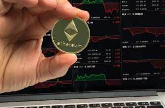 A hand holds an Ethereum coin in front of a laptop displaying cryptocurrency trading charts.