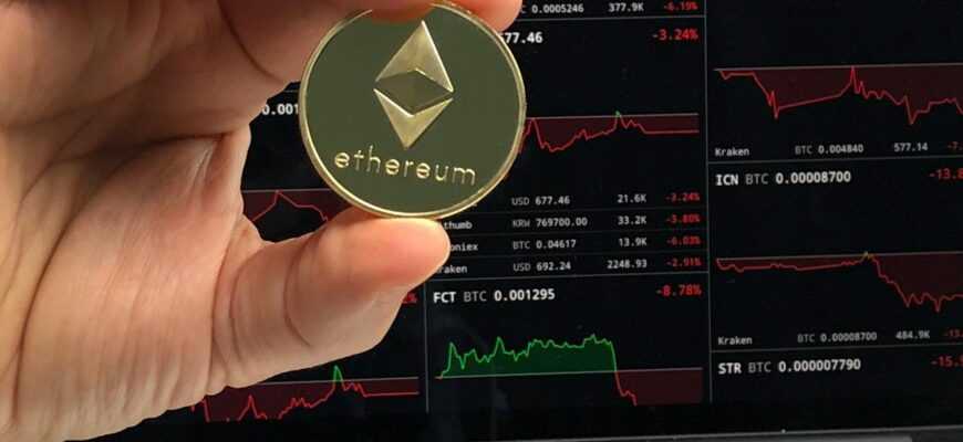 A hand holds an Ethereum coin in front of a laptop displaying cryptocurrency trading charts.