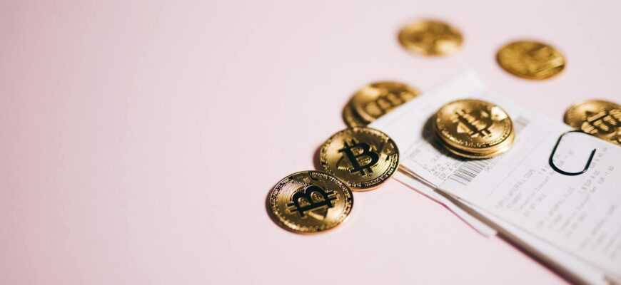 Close-up of Bitcoin coins and cash receipts on a soft pink background.